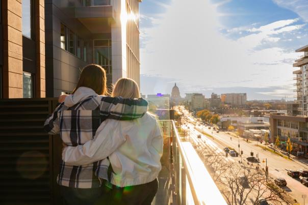 Two women stand on a balcony, overlooking a cityscape bathed in sunlight. The view includes buildings and a distant dome, with a busy street below.