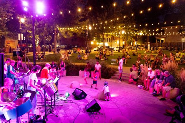 A band plays on an outdoor stage at a downtown park surrounded by lights