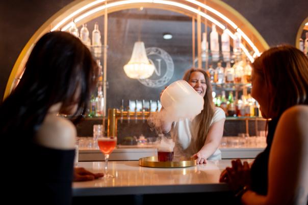 A bartender serves a cocktail with smoke effects at a stylish bar, while two patrons watch, enjoying the vibrant atmosphere.