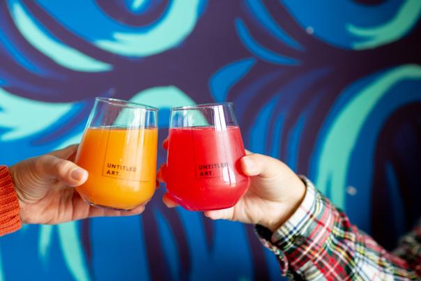 Two glasses that say "untitled art" cheersing in front of a blue and green painted wall.