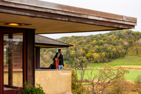 Two people stand on a covered balcony overlooking green fields and tree‑covered hills with fall foliage.