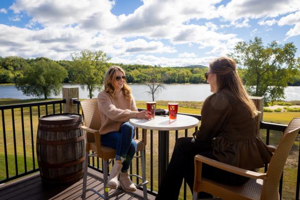 Two women sit on a deck and enjoy beers with a lake in the backghroudn