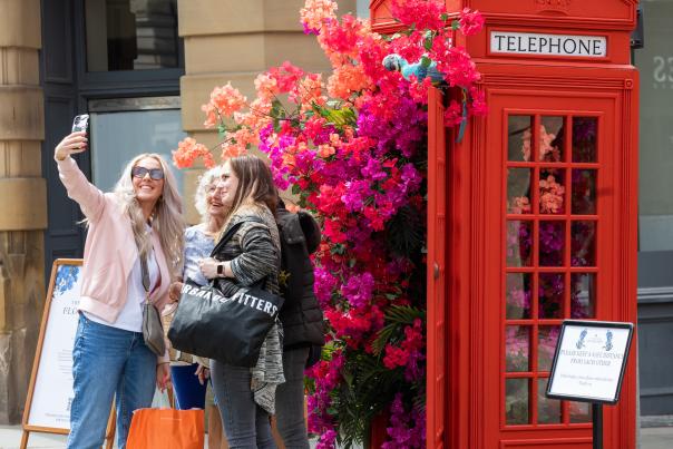 A group of women taking a selfie outside of a red telephone box with brightly coloured flowers coming out of it for Manchester Flower Festival.