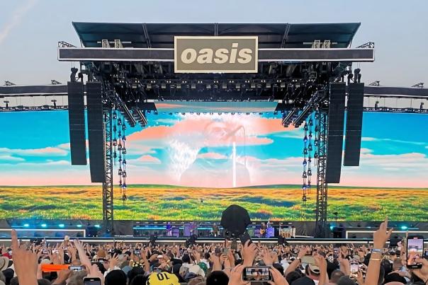 Oasis performing live on a large outdoor stage, with a crowd of fans in the foreground holding up phones and hands. The stage features a large “oasis” sign at the top, with a wide screen behind the band showing a bright landscape scene with blue sky and fields. Speakers and lighting rigs frame the stage on both sides, while the audience fills the lower half of the image, facing towards the performance.