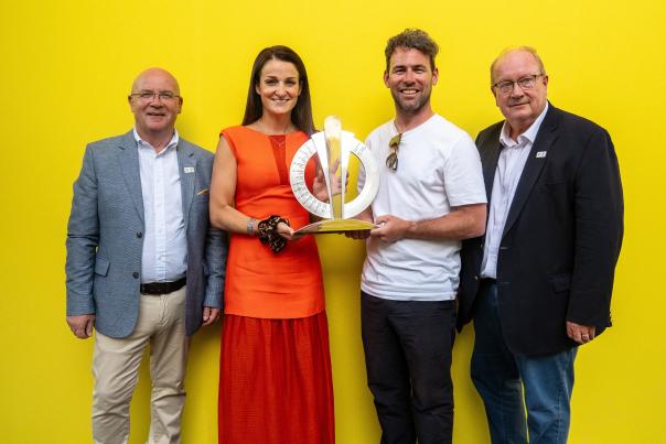 Four people stand in front of a bright yellow backdrop holding the Tour de France Grand Départ trophy during an official handover ceremony. One person in an orange outfit and another in a white shirt hold the trophy, while two others stand either side.