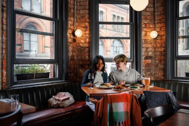 Two people seated at a table in a cosy restaurant with exposed brick walls and large windows, sharing food and drinks. Historic city centre buildings can be seen through the windows, while warm lighting and leather booth seating create an intimate dining atmosphere.