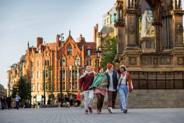 Three people walk across Albert Square in Manchester beside the ornate Manchester Cenotaph, with historic city centre buildings lining the street in the background under a clear sky.