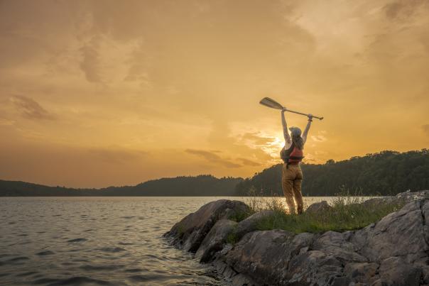 A woman raising her kayak oar the sky in delight, surrounded by a calm lake, green foliage in the distance and an orange sunset in the sky.
