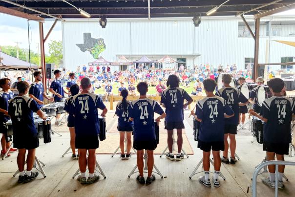 image of the backs of drumline from a DCI corps at TUPPS, looking out to the audience seated on the lawn with a white building behind them.