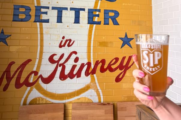 Full beer glass held in front of a mural that says Better in McKinney