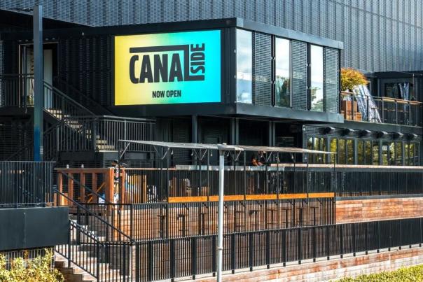 Canalside at Co-op Live in Manchester, showing a modern building with large Canal Street signage, metal railings, brick walkways and greenery alongside the canal.