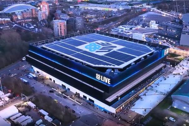 Aerial view of the Co-op Live arena in Manchester at dusk, showing solar panels on the roof and people gathering outside the venue.