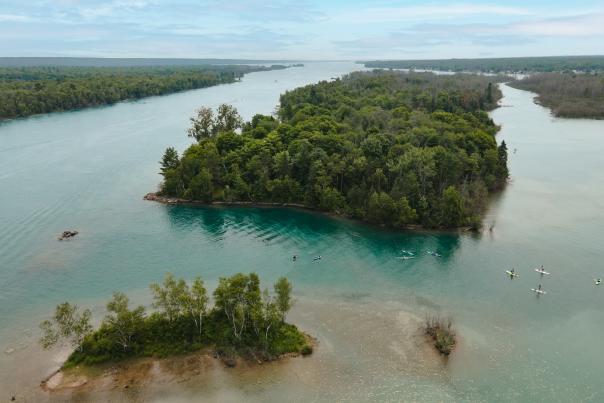 Aerial view of a lush, green island surrounded by calm blue waters, with several kayakers paddling nearby under a clear sky. Peaceful and scenic.
