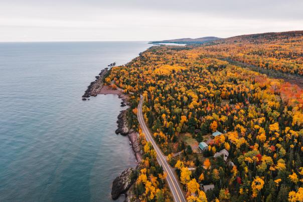 Aerial view of a winding coastal road beside a rocky lakeshore, surrounded by brilliant red, orange, and yellow autumn forest under cloudy skies.