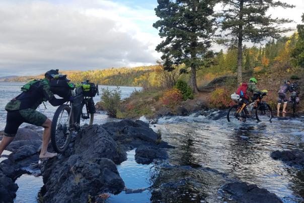 Image of mountain bikers walking bikes through rocks and water, part of "Project Adventrus" located in Michigan's Upper Peninsula