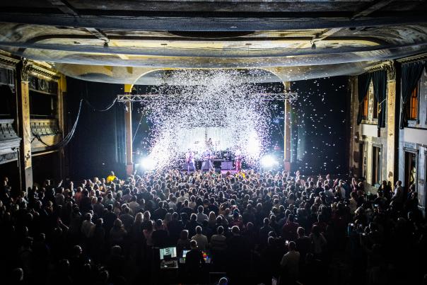 Interior view of Turner Hall ballroom during a live concert, showing a packed crowd of hundreds of people facing an illuminated stage. The historic venue's ornate architecture is visible with decorative columns, balconies, and detailed ceiling work. Dramatic stage lighting creates bright white beams cutting through the dark space, with confetti or snow effects falling from above.