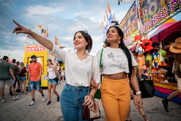 Two young women walk arm-in-arm through the Wisconsin State Fair, with one pointing excitedly ahead. Carnival rides, game booths, and crowds create a festive atmosphere in the background.