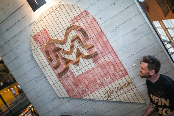 A man looks up smiling at a large wooden art piece mounted on a concrete wall. The artwork features a stylized “M” design in carved wood against a pink and white striped background, with the signature “Ike Wynter” at the bottom.