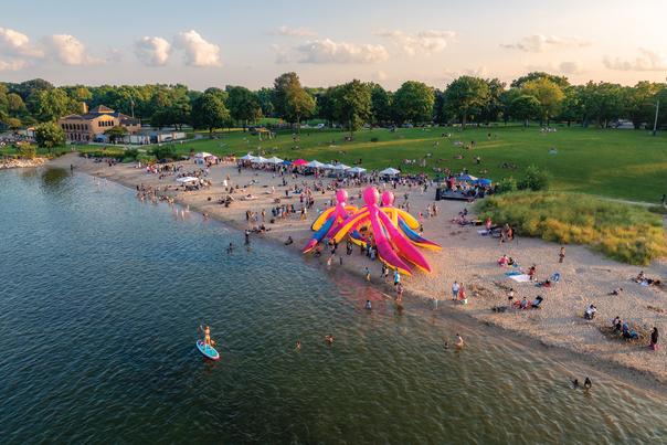 Aerial view of a vibrant beach festival along Milwaukee’s lakefront, with crowds gathered around white vendor tents and giant inflatable octopus sculptures in pink, yellow and blue. People are scattered across the sand and grass, with others swimming and paddleboarding in the lake. The scene captures a sunny day filled with art, activity and community.