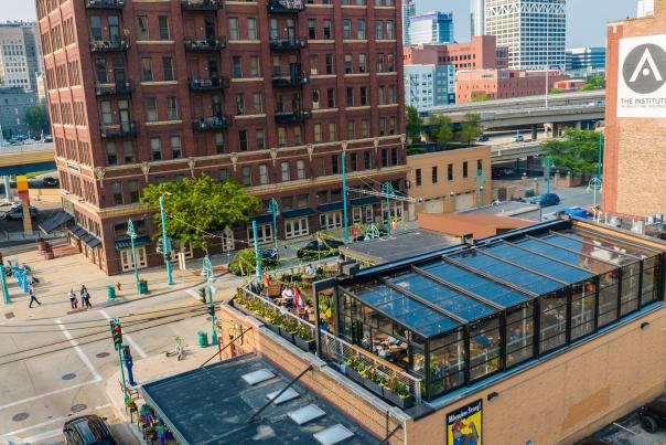 The Lux Skyhaus at Cafe Benelux, a glass greenhouse rooftop dining space in Milwaukee, overlooks a pedestrian plaza with teal lamp posts. The transparent structure reveals lush greenery and diners inside, surrounded by historic red brick buildings and downtown Milwaukee's skyline.