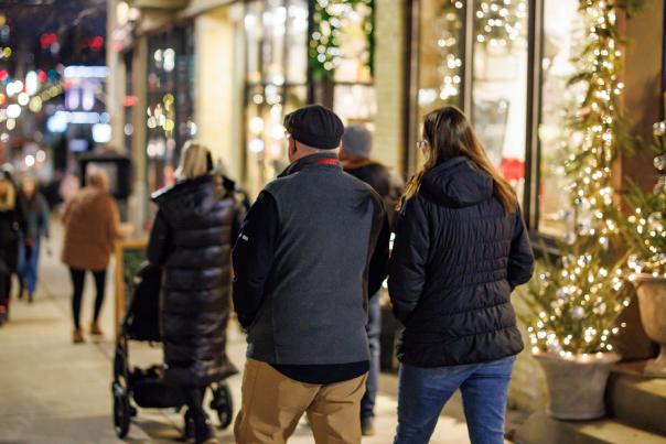 People walk along a city sidewalk decorated with holiday lights and storefront displays at night