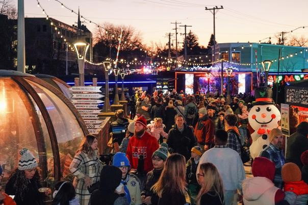 A large crowd attends an outdoor winter festival at dusk, with string lights, food tents, a clear dome shelter, and a large inflatable snowman