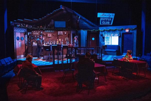 Actors perform on stage in a cozy, dimly lit theater set designed as a small-town supper club, with audience members seated at tables close to the stage for an intimate viewing experience.
