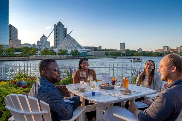 “Four friends dining outdoors at Harbor House in Milwaukee, enjoying drinks and seafood by Lake Michigan with the Milwaukee Art Museum and city skyline in the background.