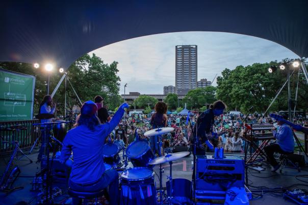 A photo taken from behind a group of musicians performing on a stage for Jazz in the Park to a crowd in Cathedral Square Park