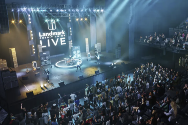 Wide view of a live concert inside Landmark Credit Union Live, with a band performing on a brightly lit stage and a large crowd gathered on the main floor and balcony seating.