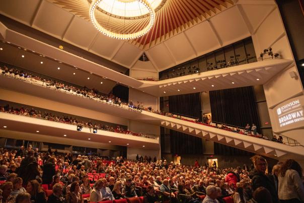 Interior of a crowded theater with multiple balcony levels, red seats, and a distinctive circular ceiling with ring chandelier