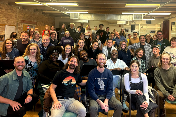The cast of Skylight Music Theatre's Joesph and the Amazing Technicolor Dreamcoat gathered in a room with brick walls and exposed wooden beams. They are seated and standing close together, posing for a group photo. The atmosphere appears friendly and cheerful, suggesting a team meeting, rehearsal, or workshop setting.