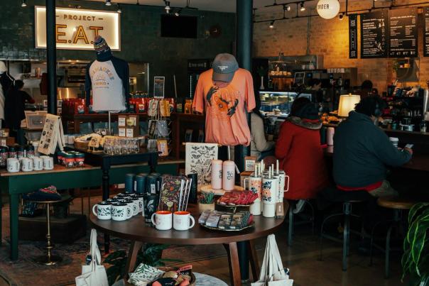 Interior of a trendy café with a round display table of merchandise like mugs, shirts, and tote bags in the foreground, and people seated at the counter enjoying drinks in the background.