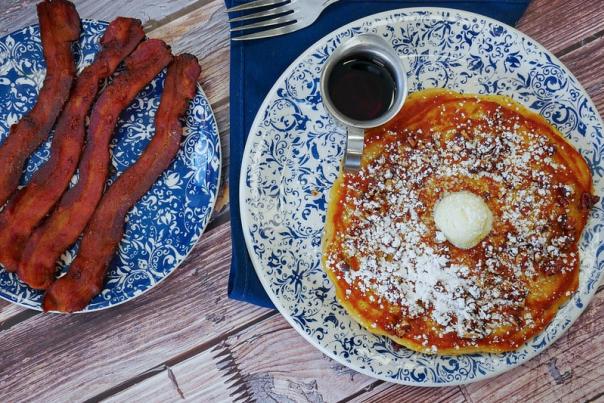 A decorative blue-and-white plate holds a sweet potato pancake topped with powdered sugar, a pat of butter, and a small metal cup of syrup on the side. Next to it, another matching plate features several strips of crispy bacon. A fork rests on a dark blue napkin beside the pancake plate, all set on a rustic wooden table.