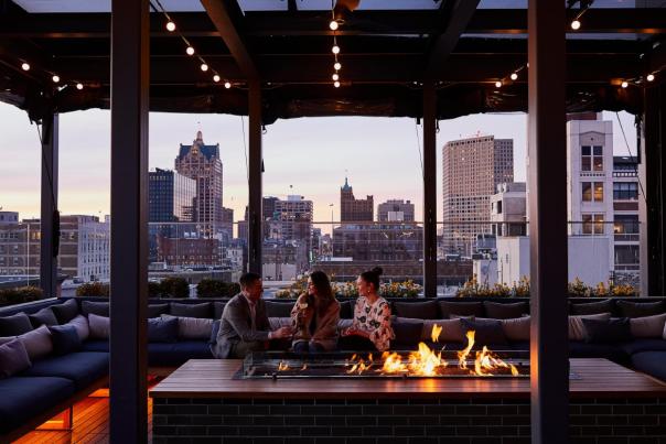 Three people sitting around a rooftop fire pit at sunset, enjoying drinks with a view of downtown Milwaukee’s skyline at The Outsider rooftop bar.