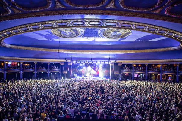 A packed concert crowd fills the ornate ballroom of The Rave/Eagles Club in Milwaukee, Wisconsin. Bright stage lights illuminate the performers, while balconies and a large, domed ceiling with gold detailing frame the historic venue.