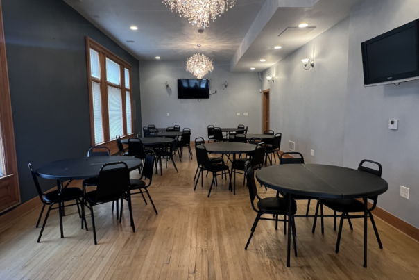 A bright event room with hardwood floors, round tables and black chairs, two wall-mounted TVs, crystal chandeliers, and large windows along one wall.