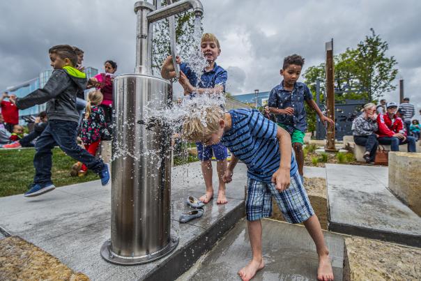 Children play joyfully at an outdoor water pump, one boy splashes under the water, others giggle nearby. Cloudy sky and spectators in the background.
