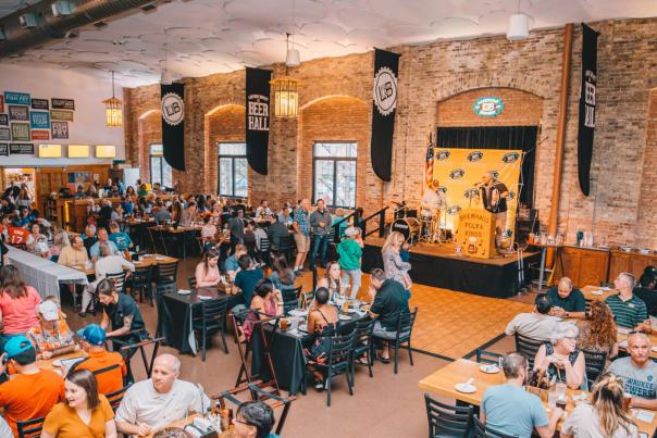 An open banquet hall filled with people eating food and drinking beer while a performer plays the accordion in the background.