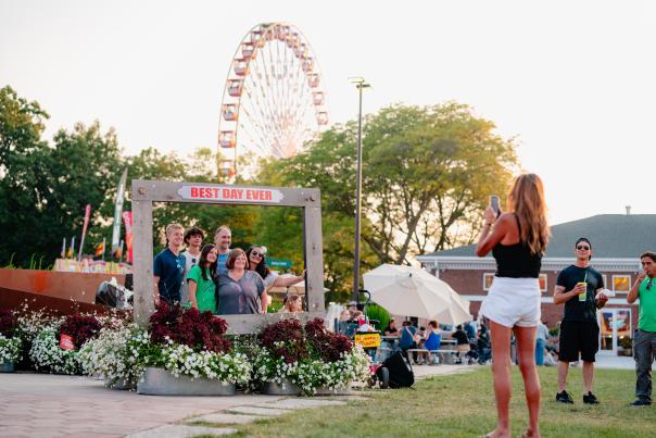 A woman photographs five people posing inside a wooden “Best Day Ever” frame surrounded by flowers, with the Ferris wheel rising above the trees at the Wisconsin State Fair.