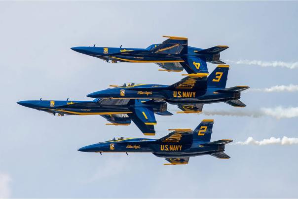 Four U.S. Navy Blue Angels fighter jets fly in tight formation against a clear sky, showcasing precision and skill during an aerial demonstration. The jets are painted in blue and yellow, trailing faint white smoke.