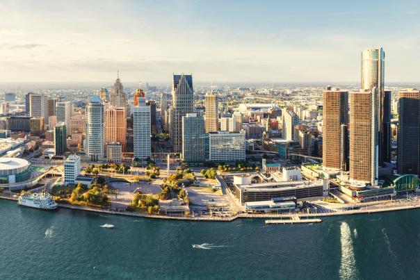 Aerial view of downtown Detroit along the Detroit River, with the city skyline, waterfront buildings and boats visible in the foreground.