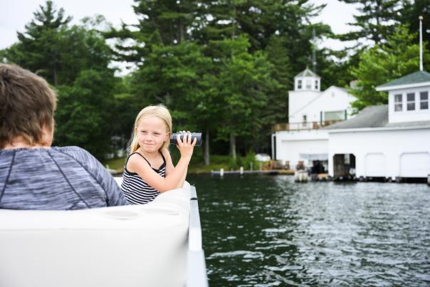Little girl sitting on a pontoon and looking through binoculars at a white boathouse on the lake.