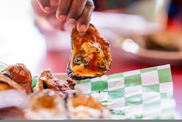 A hand holds a grilled oyster topped with herbs, above a plate of oysters on a green and white checkered paper. A vibrant dining setting is visible.