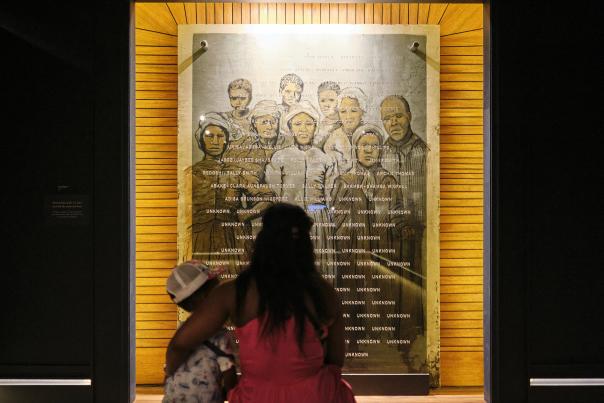 A Black woman with long hair holds her child while they look at a photo collage of formerly enslaved residents of Africatown, Alabama