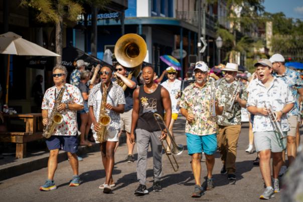 A band of five men with various musical horns are dressed in hawaiian shirts playing music while walking down the street.