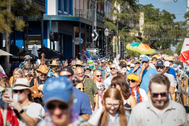 A large group of people in colorful clothing, hats and umbrellas walk down the street between buildings for a parade