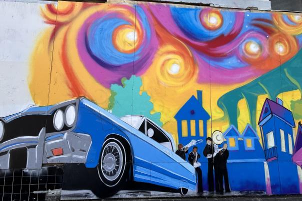 Mural of blue car in front of a rainbow sky and cityscape