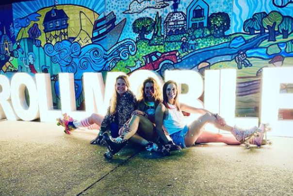 three women sitting on a sidewalk in front of 3 foot lighted letters reading # roll mobile