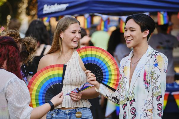 Two people laughing and smiling while holding rainbow colored folding fans.